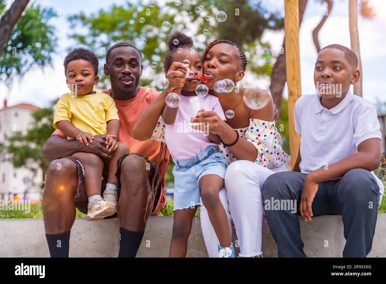 African black ethnic family with children in playground blowing soap bubbles next to trees in the park Stock Photo