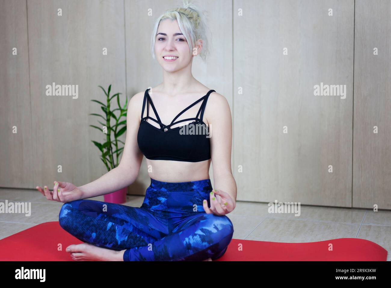 People, meditation and relaxation. Attractive smiling blonde woman wearing top and trousers sitting on fitness mat, Stock Photo