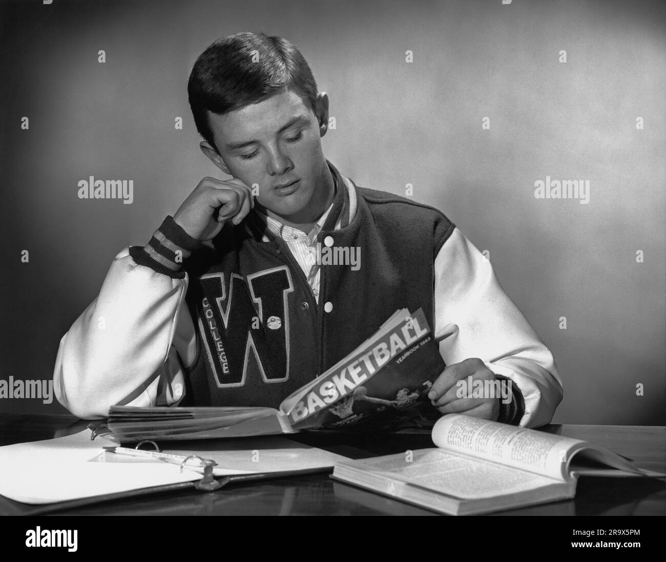 Teenage college boy in sports jacket with his college logo W as takes a minute and pauses from doing schoolwork to read a sports/basketball magazine Stock Photo