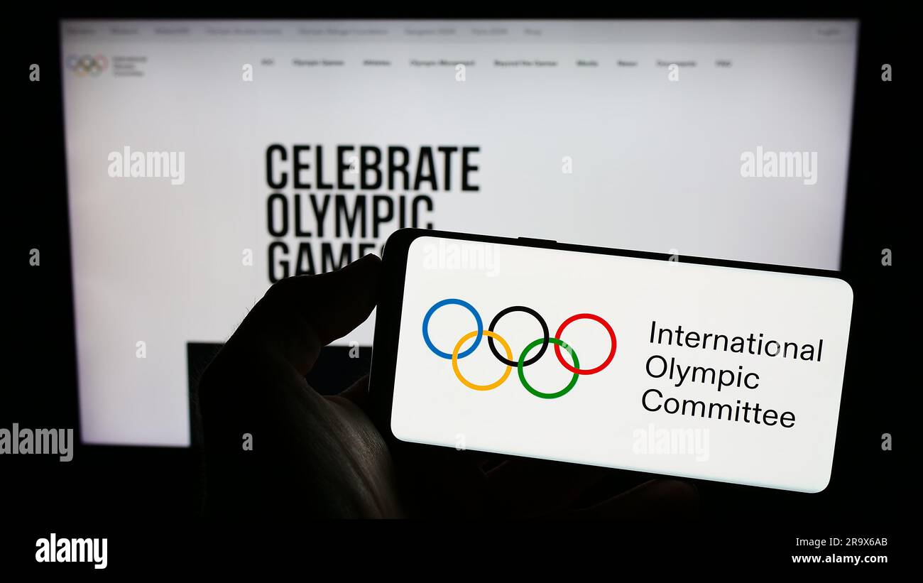 Person holding cellphone with logo of the International Olympic Committee (IOC) on screen in front of webpage. Focus on phone display. Stock Photo