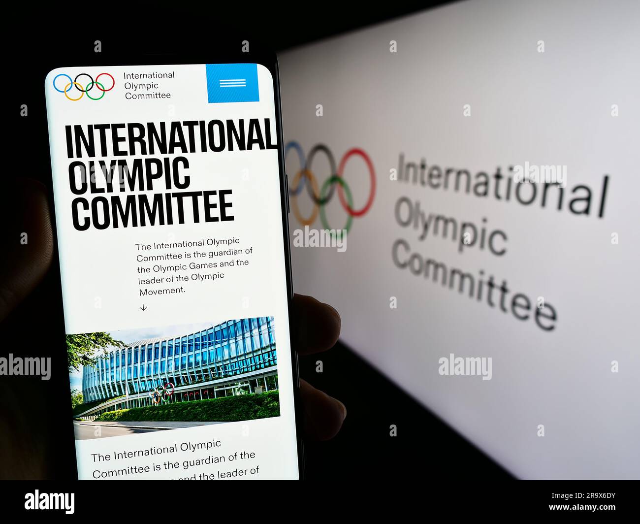 Person holding smartphone with website of the International Olympic Committee (IOC) on screen in front of logo. Focus on center of phone display. Stock Photo