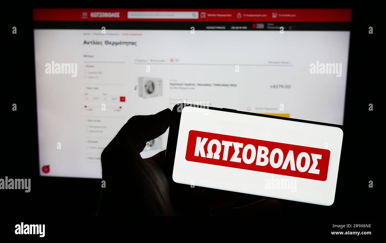 Person holding cellphone with logo of Greek electronics retail company Kotsovolos on screen in front of business webpage. Focus on phone display. Stock Photo