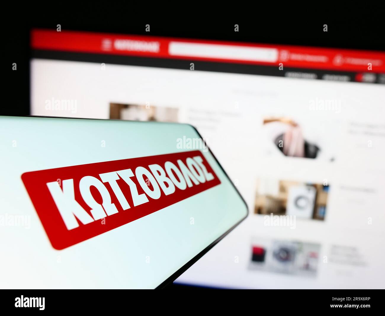 Mobile phone with logo of Greek electronics retail company Kotsovolos on screen in front of website. Focus on center-left of phone display. Stock Photo
