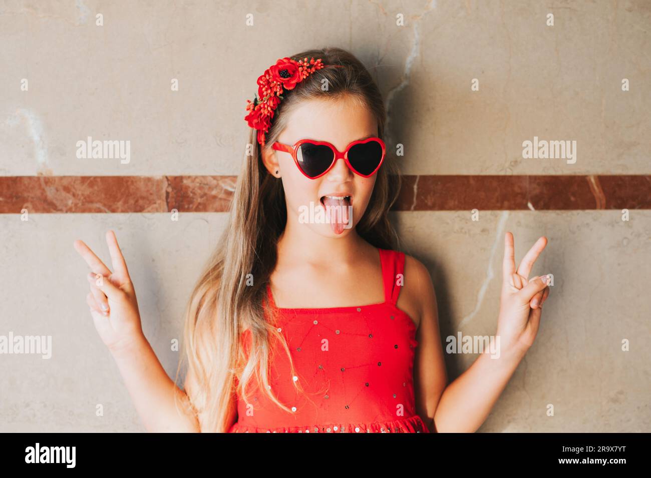 Funny portrait of pretty little girl wearing red dress and heart shaped sunglasses, pulling tongue Stock Photo