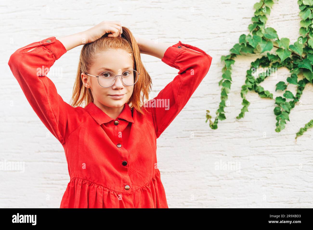 Outdoor portrait of cute preteen girl wearing red dress and eyeglasses, holding hair tail Stock Photo