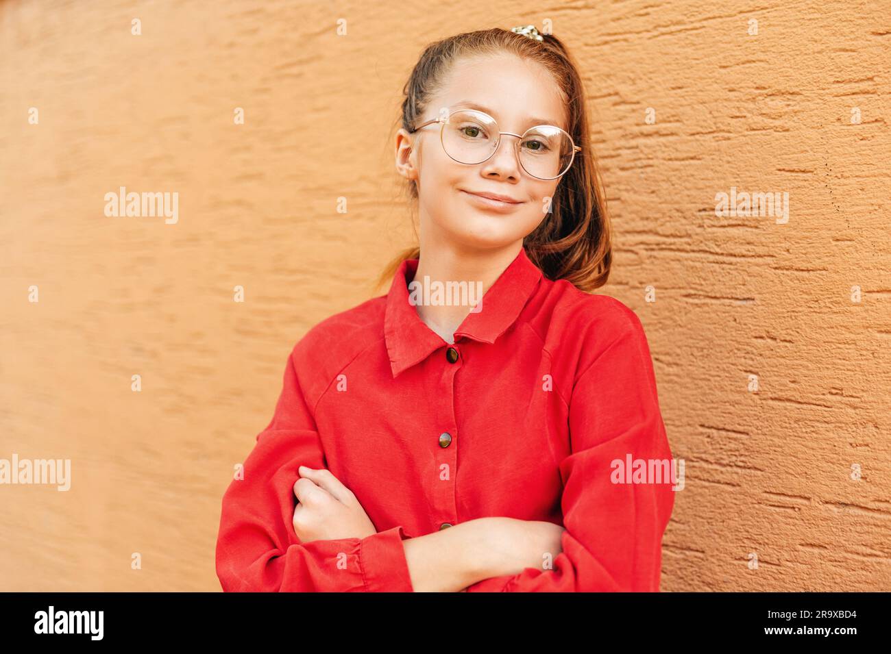 Outdoor portrait of cute preteen girl wearing red dress and eyeglasses, posing against orange wall Stock Photo