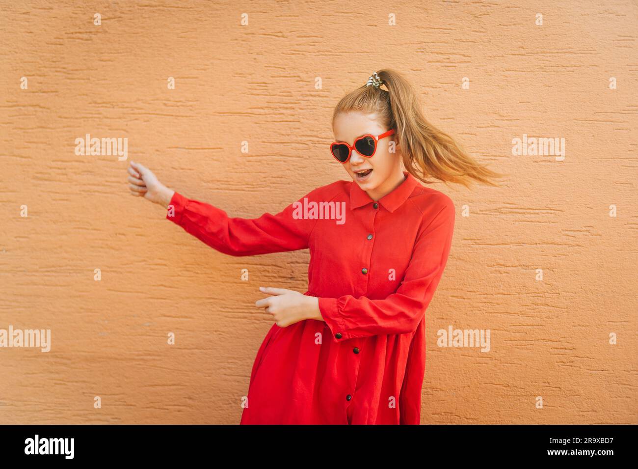 Happy dancing preteen girl, wearing red dress, heart shaped sunglasses, orange wall on background Stock Photo