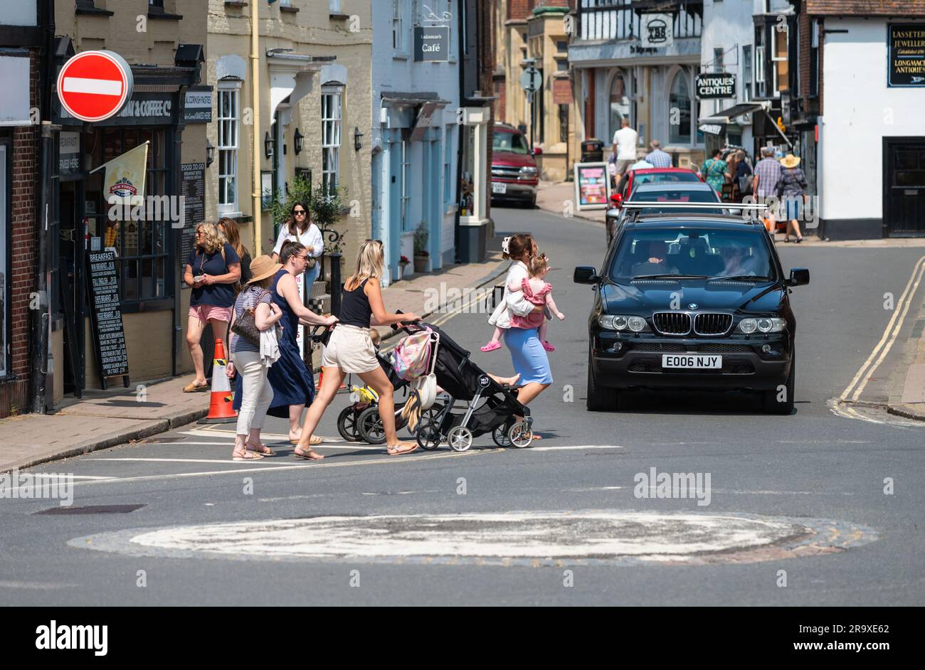 Women with children in pushchairs, presumably Mothers, pushing them across a British road while a car driver waits at a mini roundabout in England, UK Stock Photo