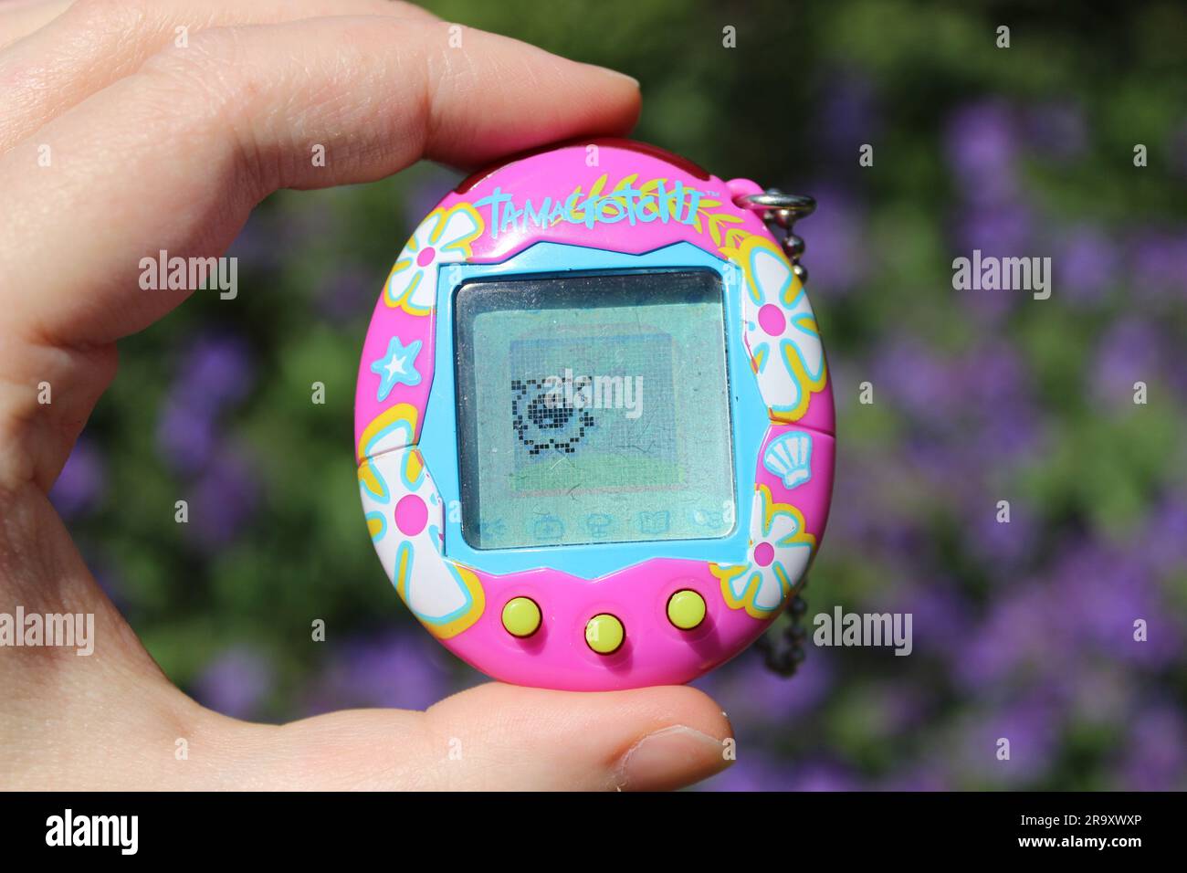 Photo depicting a tamagotchi device, pink with tropical flowers, held in someone's hand. Stock Photo