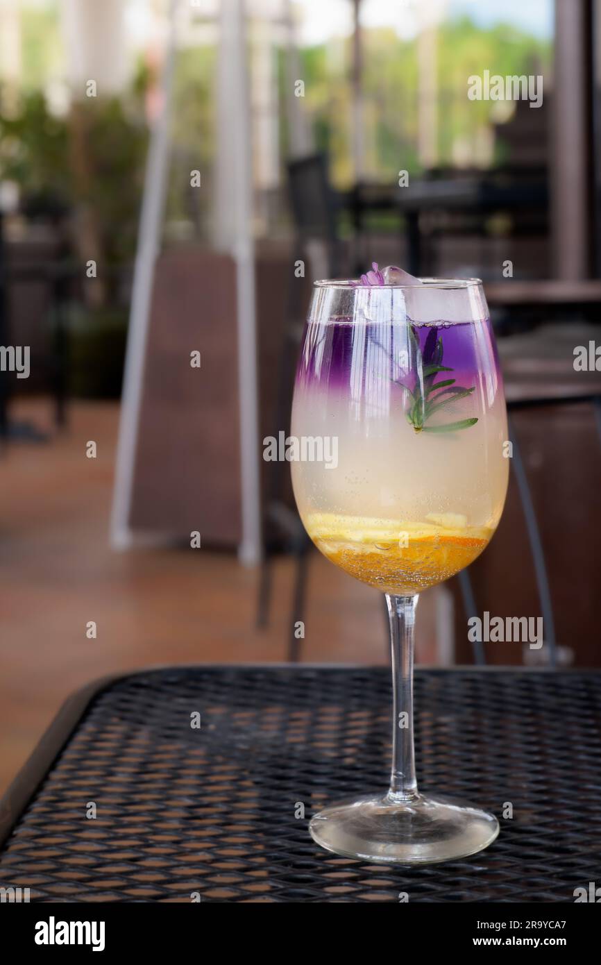 colorful gourmet craft cocktail made with purple gin garnished with a flower and orange slice with soft focus outdoor patio background Stock Photo