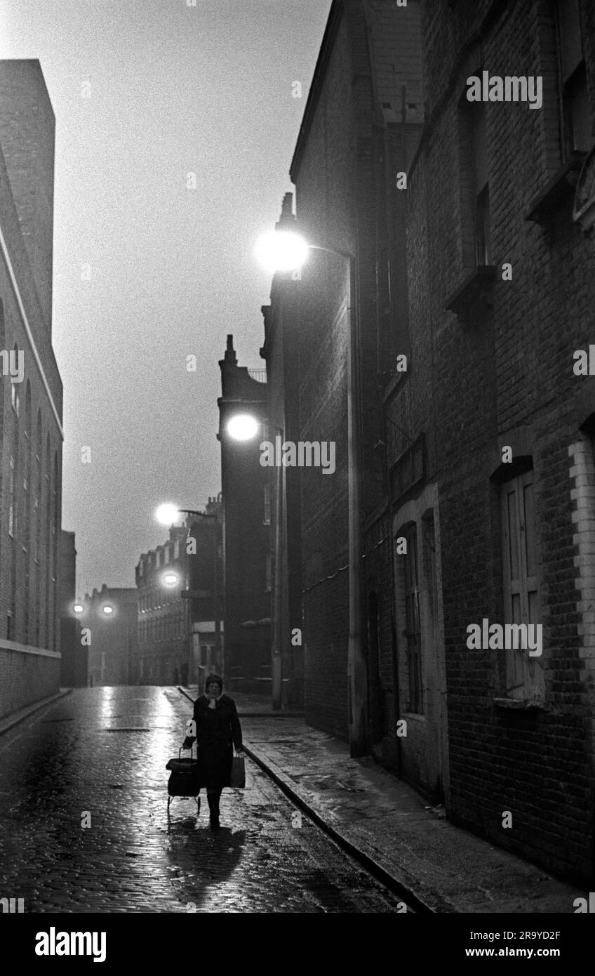 East End London 1970s UK. Brick Lane, a woman residents pulls her shopping trolly bag along the cobbled street past Victorian tenement blocks of flats. Peabody Estate Tower Hamlets, London, England 1976. HOMER SYKES Stock Photo