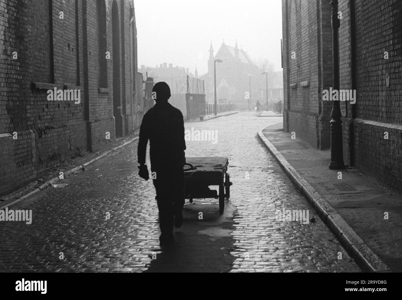 Whitechapel East London 1970s. Working class man pulls a handcart along the cobbled street past huge windowless warehouses. In the distance is St Annes Church. the area has been completely redeveloped. Tower Hamlets, London, England circa 1974 70s UK HOMER SYKES Stock Photo