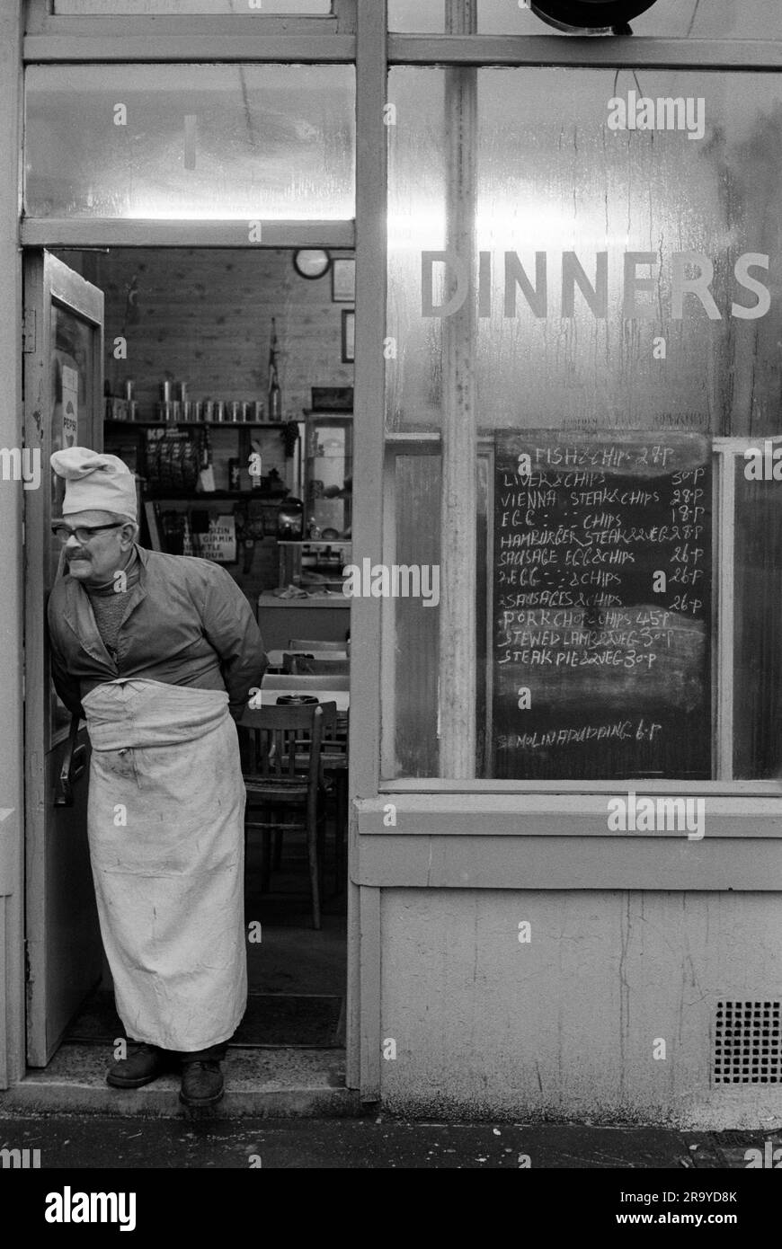 Brick Lane London 1970s. The proprietor in chefs hat and apron, buttoned up to his jacket, stands outside his greasy spoon Dinners café in Brick Lane waiting for lunchtime customers. The blackboard dinner menu is almost everything with chips. Whitechapel, London, England 1974. 70s UK HOMER SYKES Stock Photo