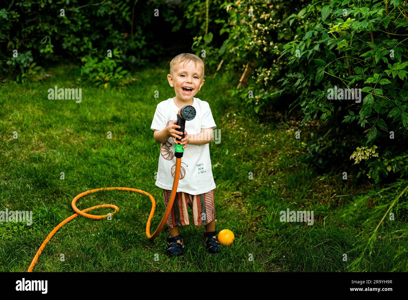 Boy watering flowers and plants in home garden. Child with water hose in summer backyard. Family activity with family. Children help parents. Stock Photo