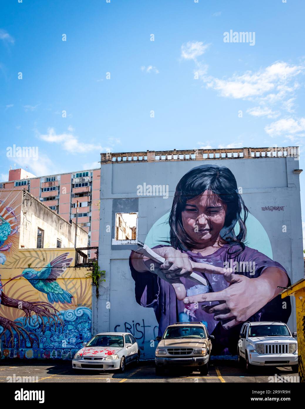 Public art mural on building in Santurce neighborhood in San Juan Puerto Rico with girl artist holding paintbrush on wall above parking lot. Stock Photo