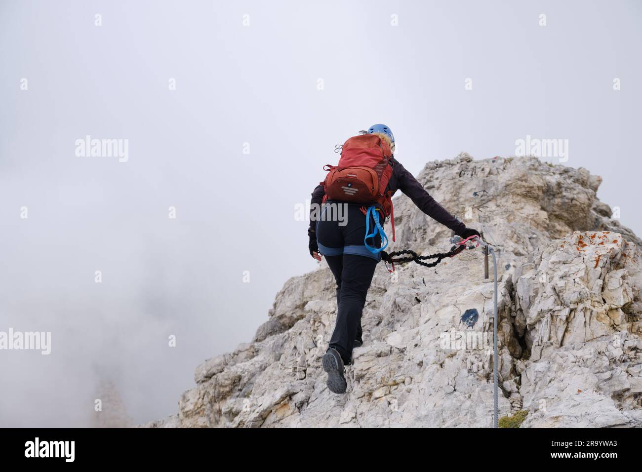 Active woman secured to via ferrata cable, advances on a route at Tofana di Mezzo, Dolomites, Italy, on a ridge surrounded by fog. Adventure, courage, Stock Photo