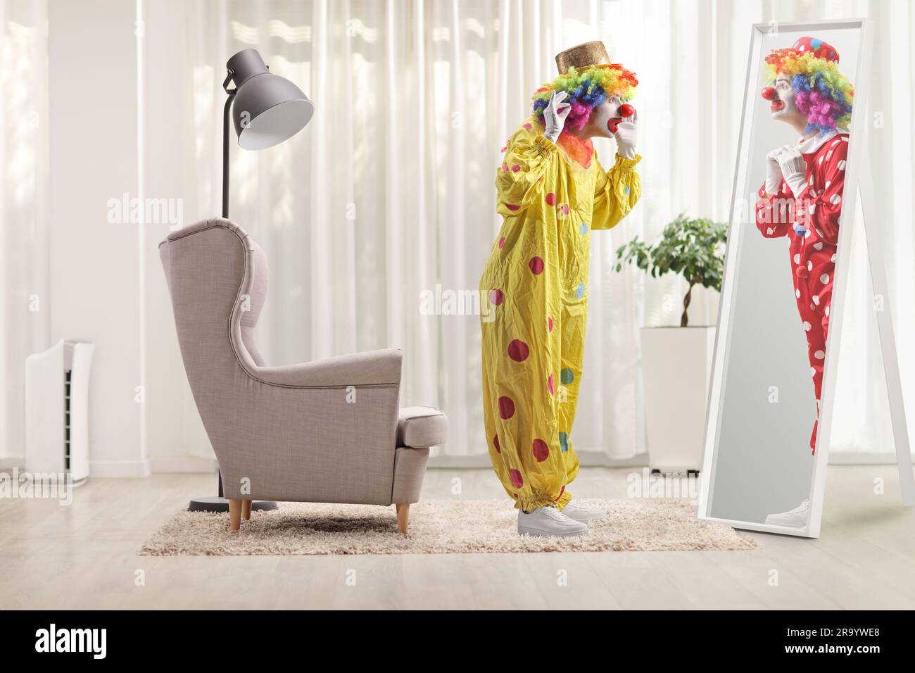Clown in a yellow costume standing in front of a mirror and looking at a clown in a red costume in a room Stock Photo