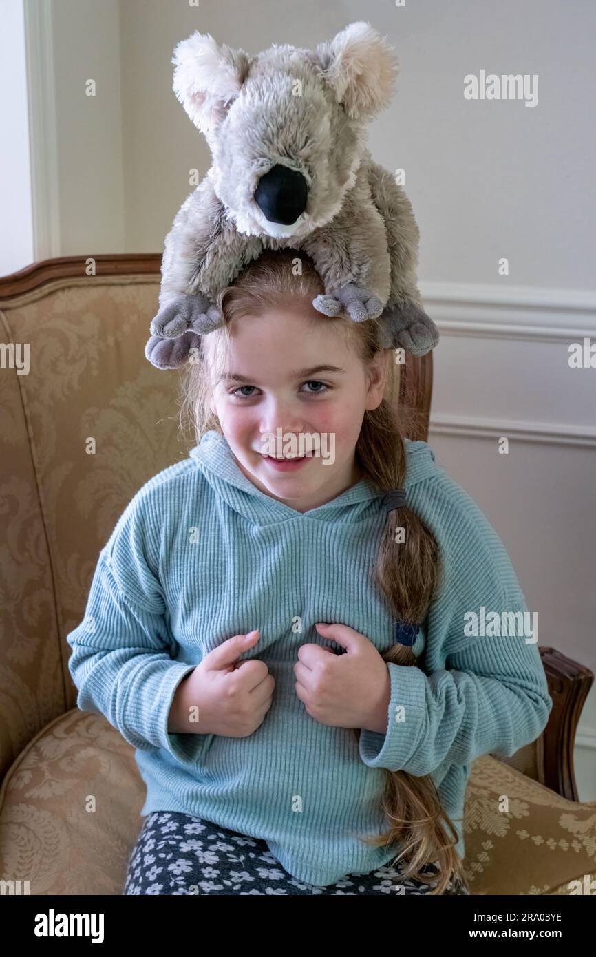 Portrait of a smiling 7 year old girl balancing a stuffed koala bear on her head.  (MR) Stock Photo