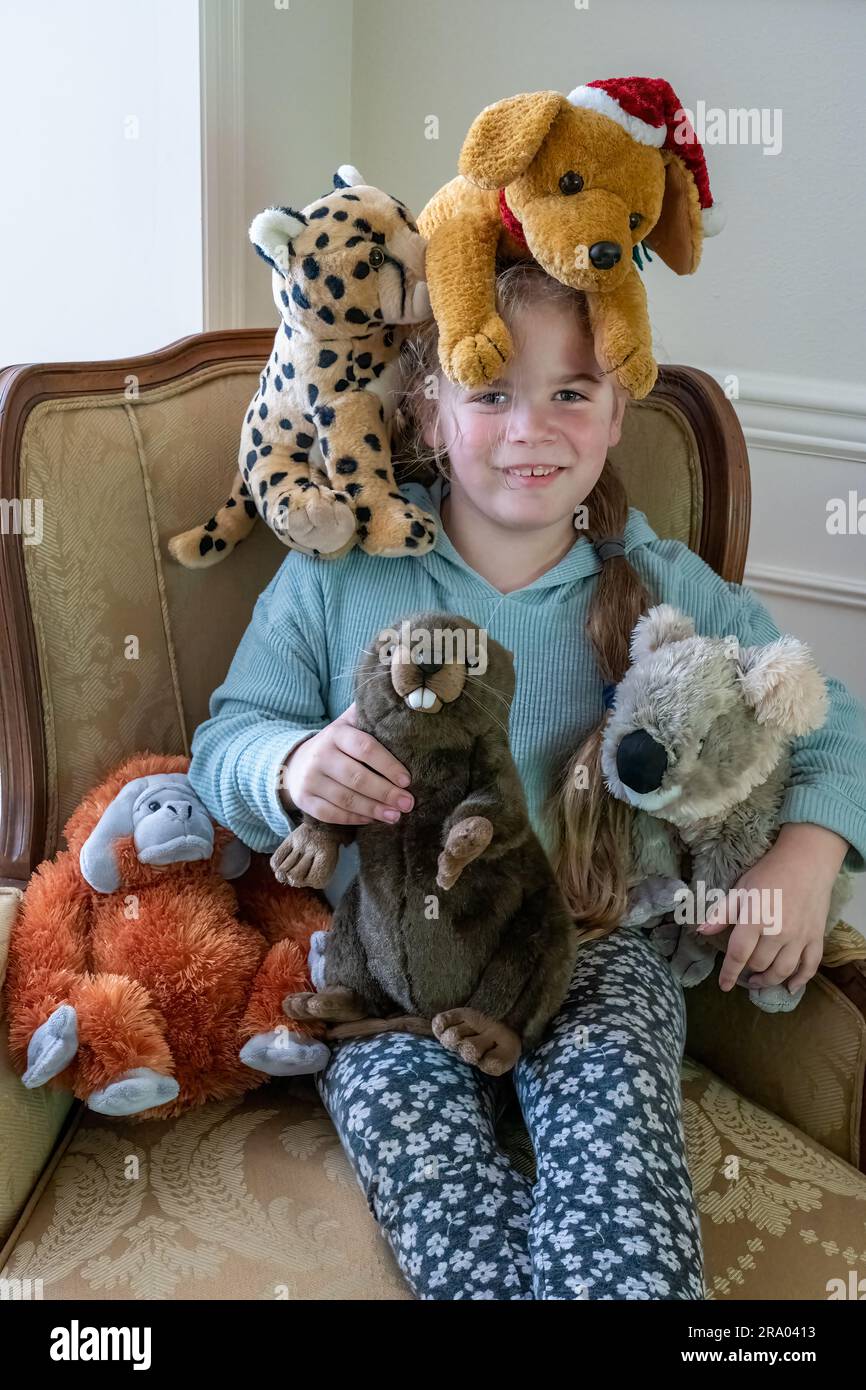 Portrait of a 7 year old girl sitting in an armchair surrounded by stuffed animals, including a dog on her head.  (MR) Stock Photo