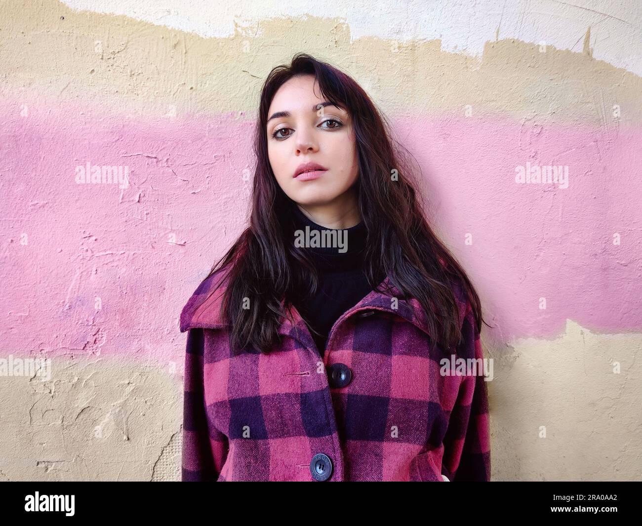 Thoughtful woman with dark hair, in black golf and in a purple plaid coat against the background of a colored wall. Thoughtful state. Stock Photo