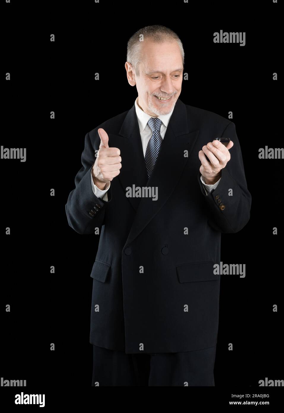An amiable businessman wearing a black suit smiling and reading on mobile phone, with thumb up, on black background Stock Photo
