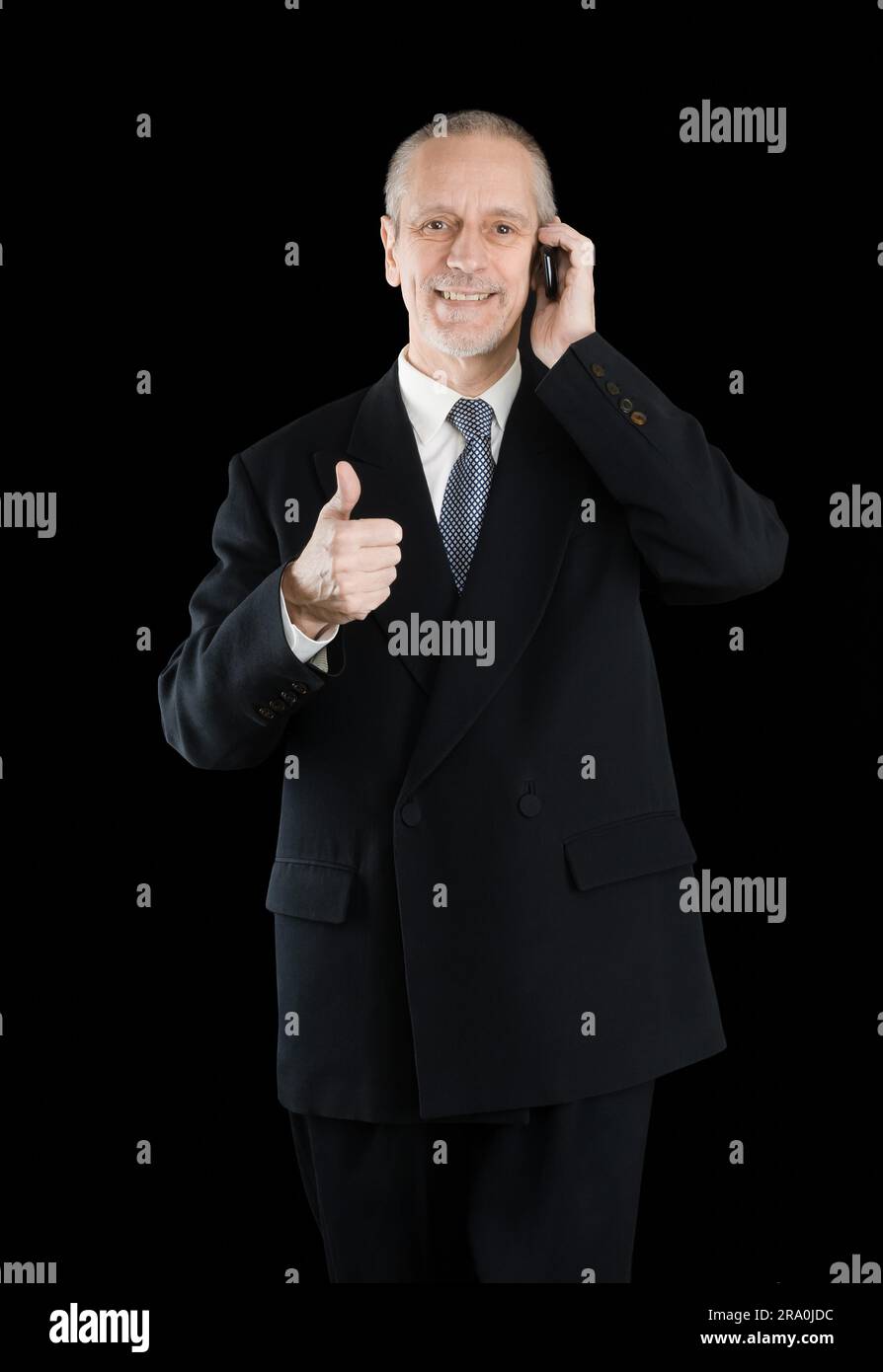An amiable businessman wearing a black suit smiling and speaking on mobile phone, with thumb up, on black background Stock Photo