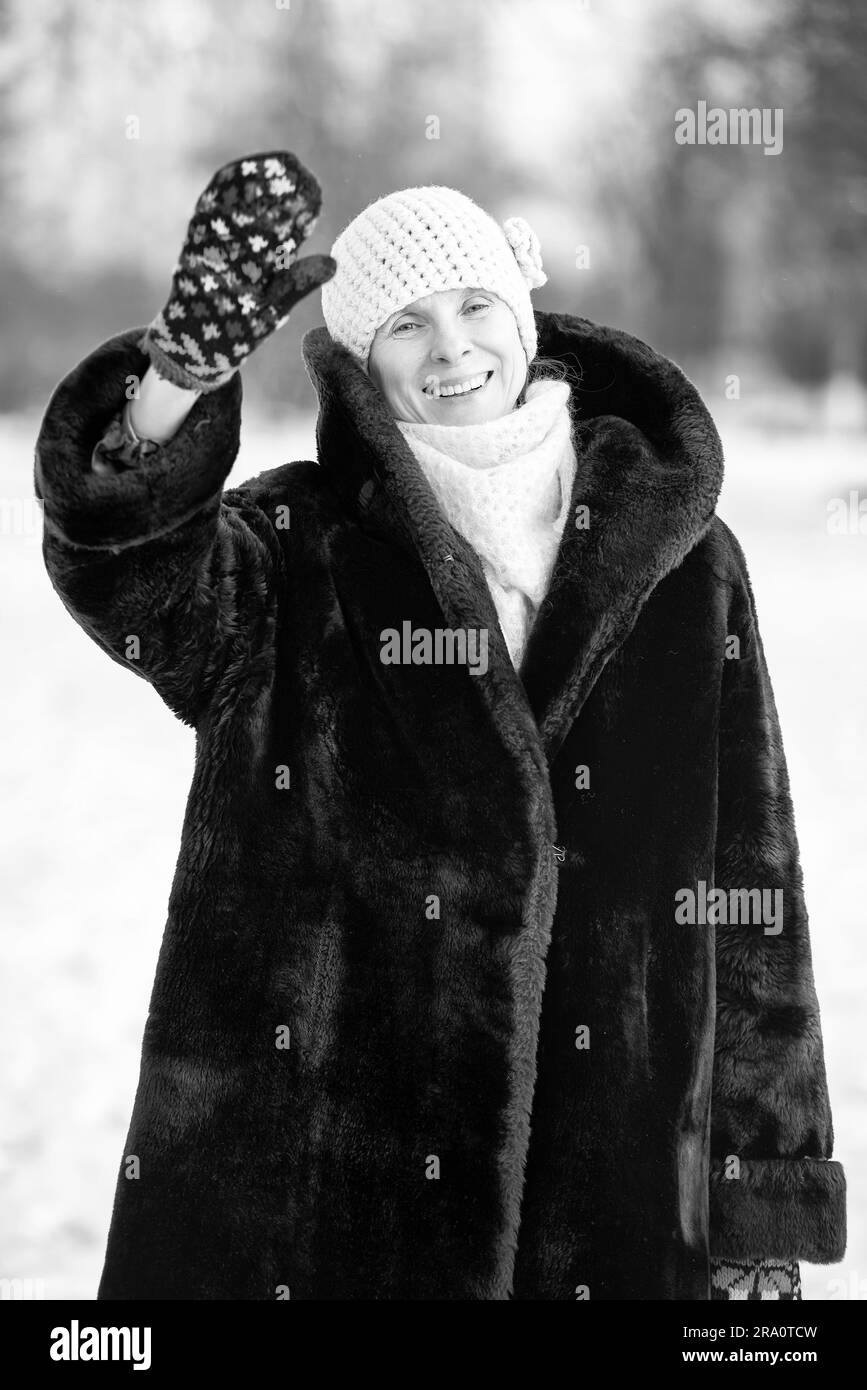 A winter portrait of a smiling senior adult woman wearing a wool cap, a scarf and colored gloves, saluting with her hand, with a snow background Stock Photo