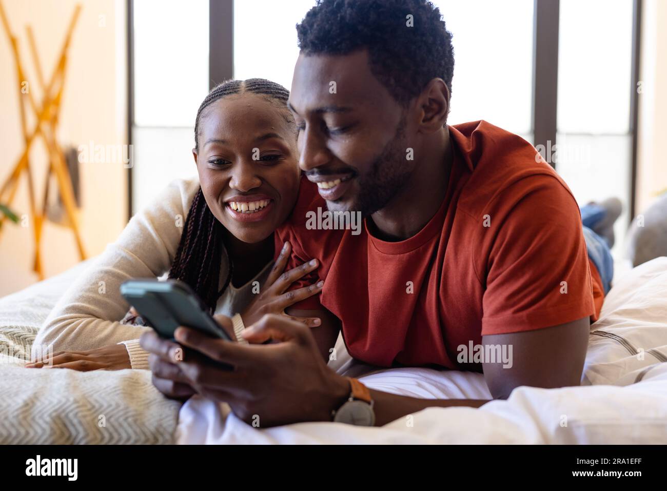 Happy african american couple using smartphone lying on bed in bedroom ...
