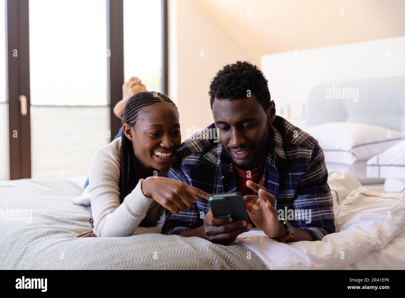 Happy african american couple using smartphone lying on bed in bedroom ...