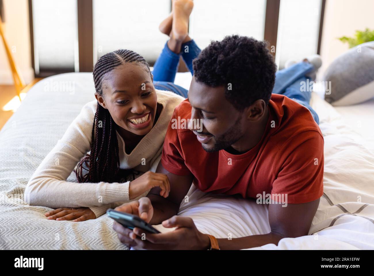 Happy african american couple using smartphone lying on bed in bedroom ...