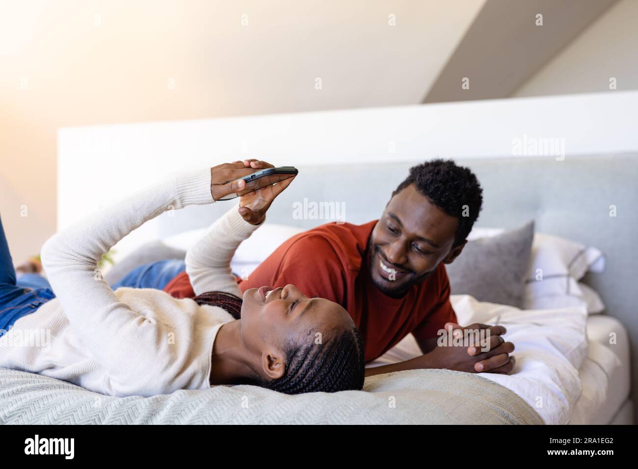 Happy african american couple using smartphone lying on bed in bedroom ...