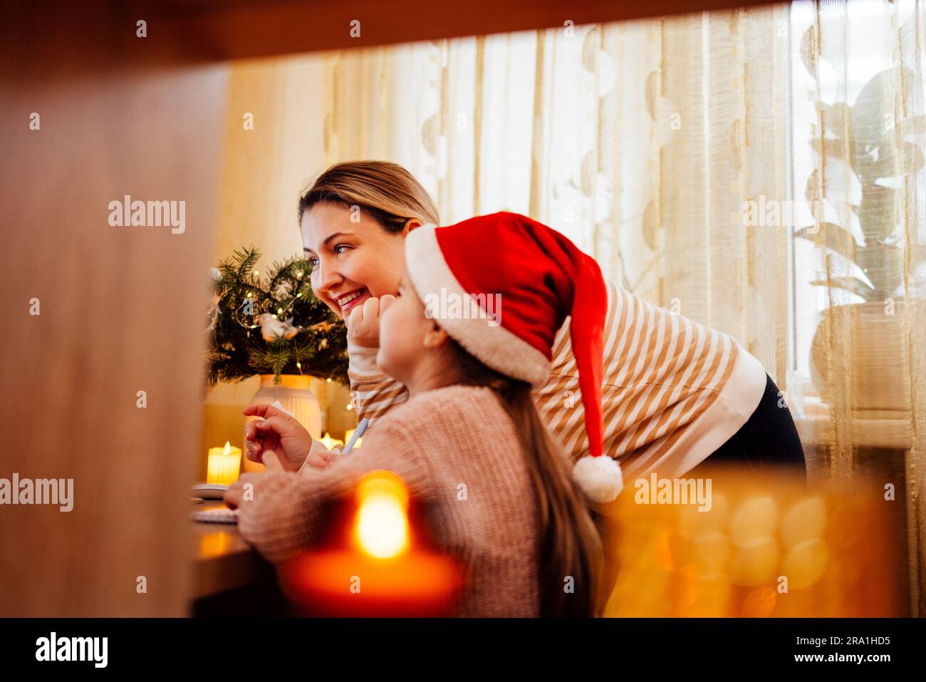 Young woman and her sweet little daughter in santa hat happily communicate via video link with friends. Smiling blonde mom and her cute kid congratula Stock Photo