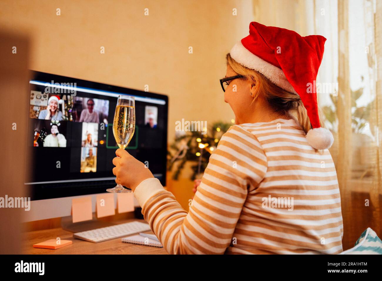 Young woman in santa hat happily communicates via video link with friends. Smiling girl holds glass of champagne and sparkler in her hands and congrat Stock Photo