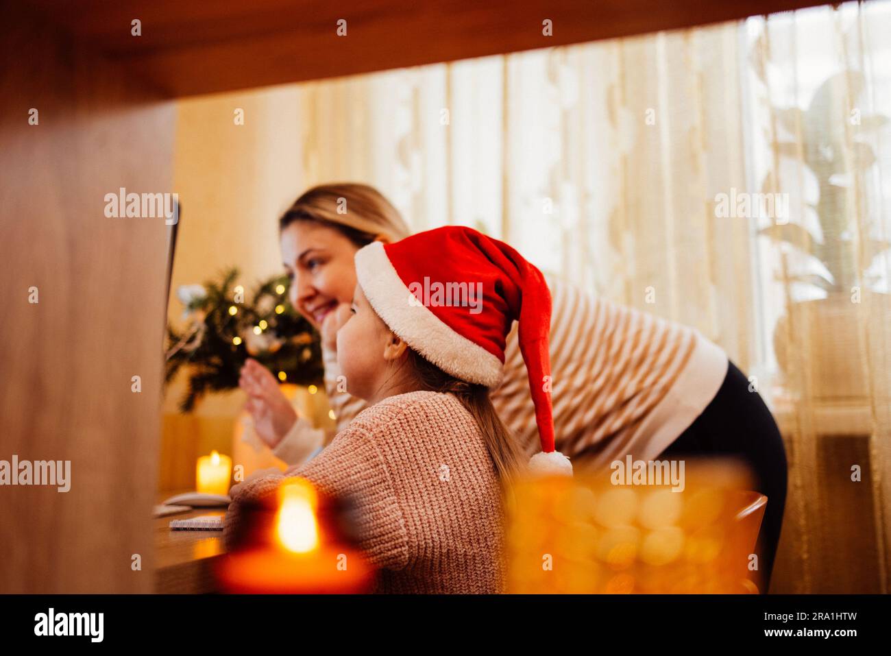 Young woman and her sweet little daughter in santa hat happily communicate via video link with friends. Smiling blonde mom and her cute kid congratula Stock Photo