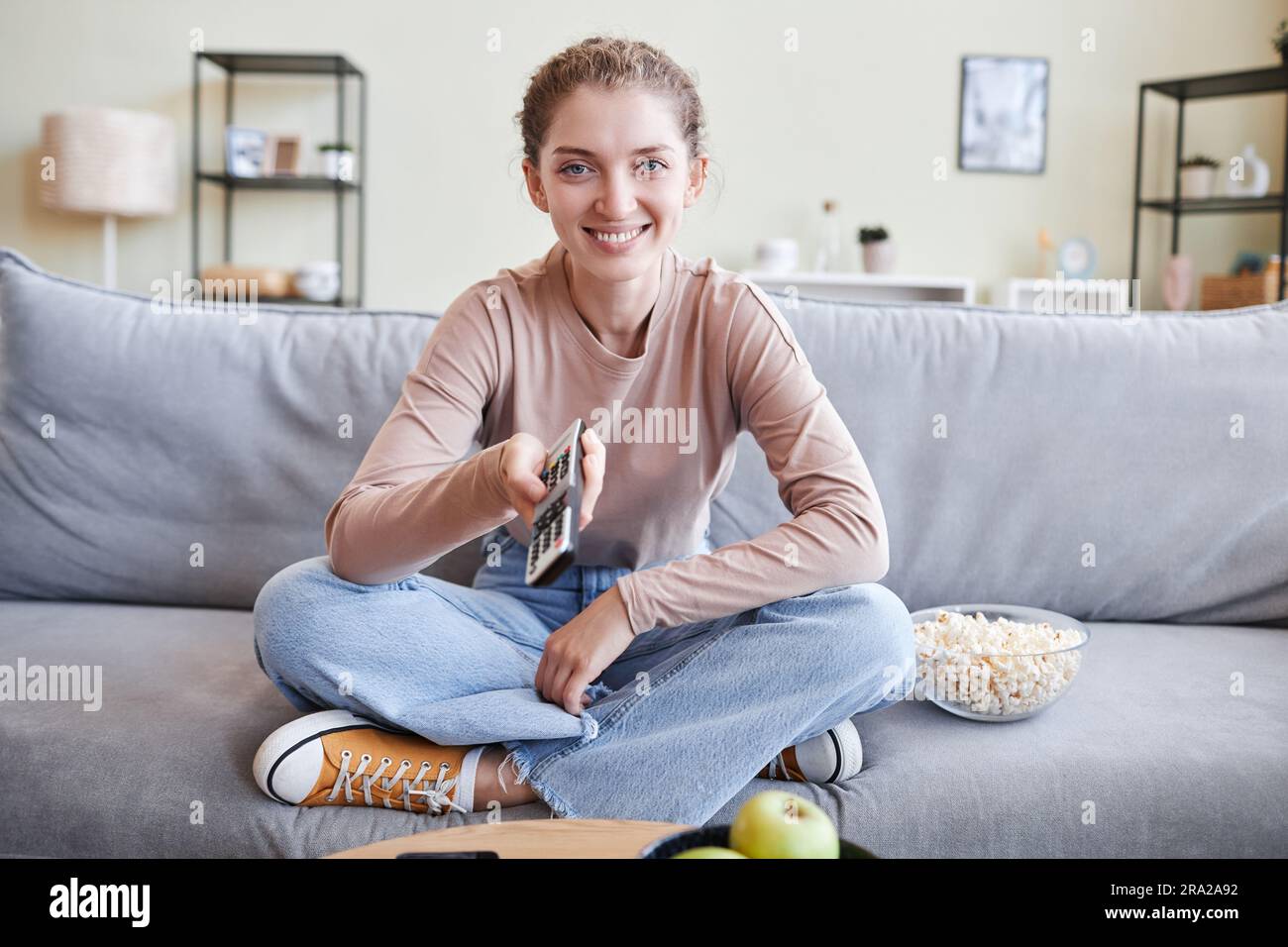 Front view portrait of carefree young woman sitting on sofa with legs crossed and holding remote control looking at camera, copy space Stock Photo
