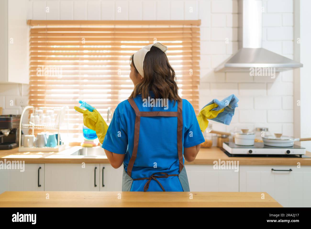 Efficient and resourceful, an Asian housewife confidently holds a tablecloth and sprayer in the kitchen, showcasing her dedication to maintaining a cl Stock Photo