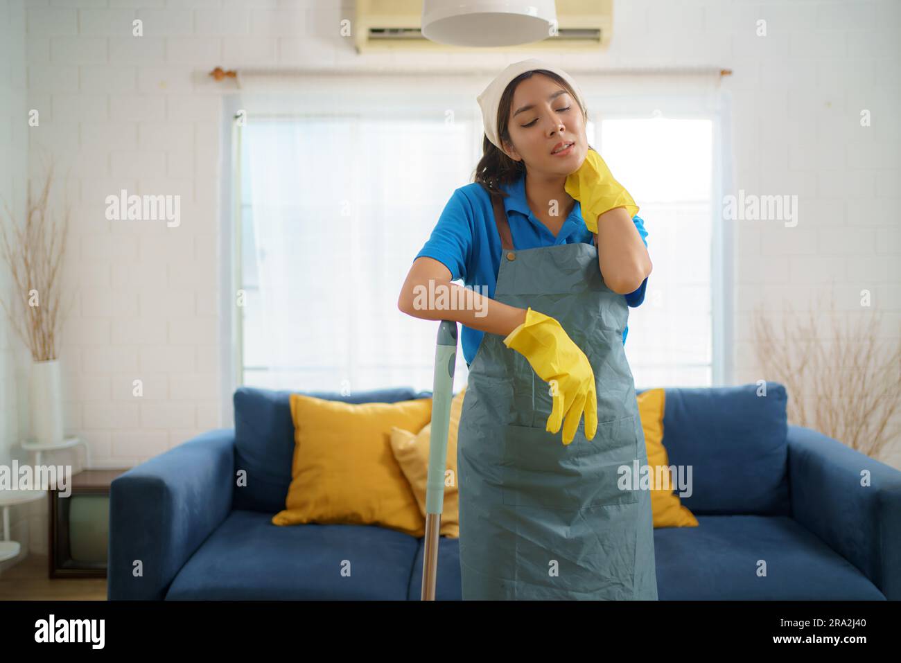 Exhausted Asian maid struggles with fatigue while mopping the house, showcasing the challenges and hard work behind domestic house Stock Photo