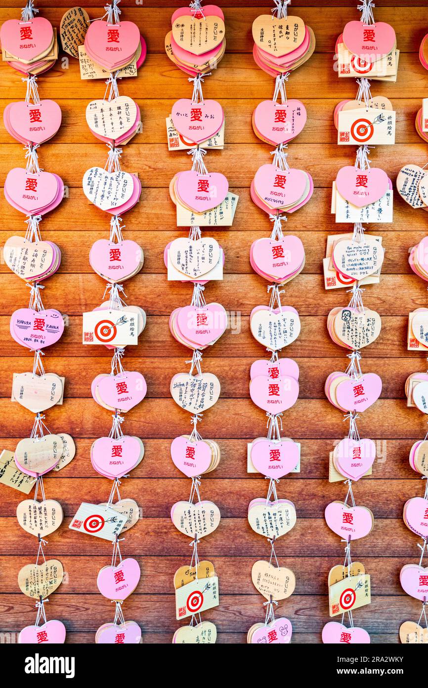 Rows of Japanese Shinto ema hanging on wooden board. Pink heart shaped wooden wishing plaques mixed with traditional shaped ones at Ikuta Shrine, Kobe Stock Photo