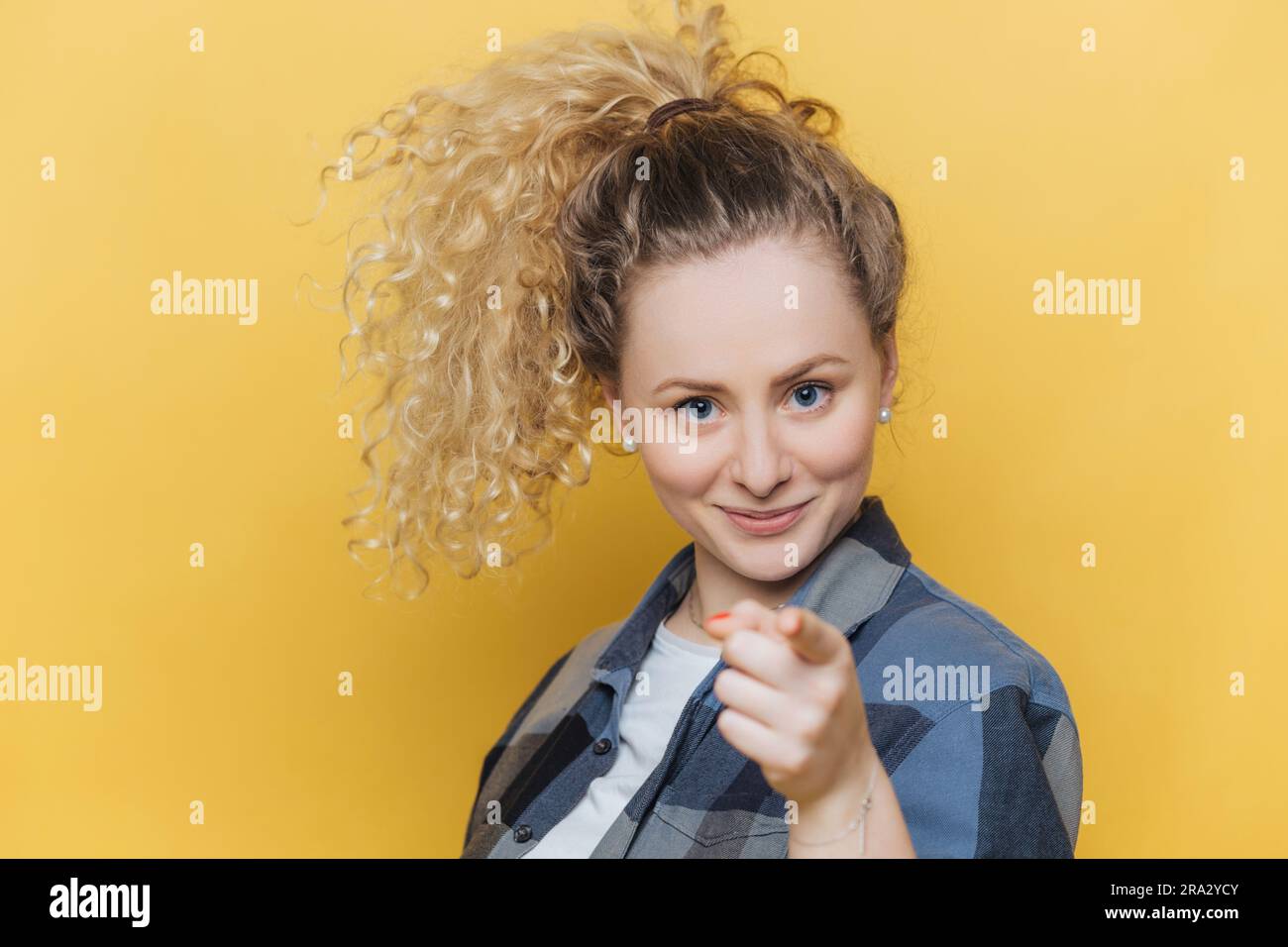 Confident woman chooses you, dressed casually, pointing at camera indoors. Stock Photo