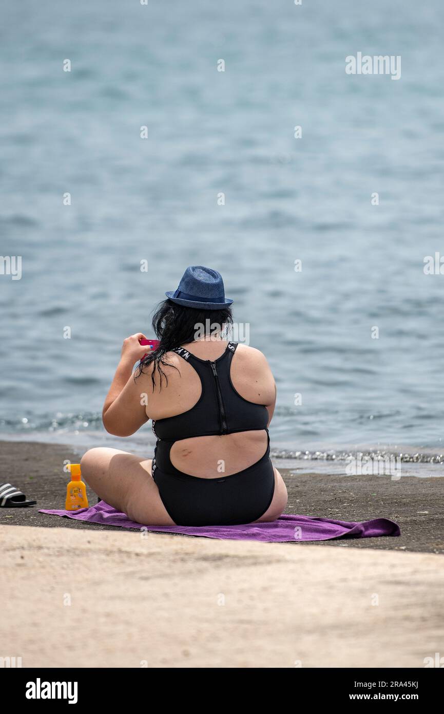 large lady wearing black swimming costume and sun hat sitting on a beach at the water edge. overweight woman seated on a towel at the seaside. Stock Photo