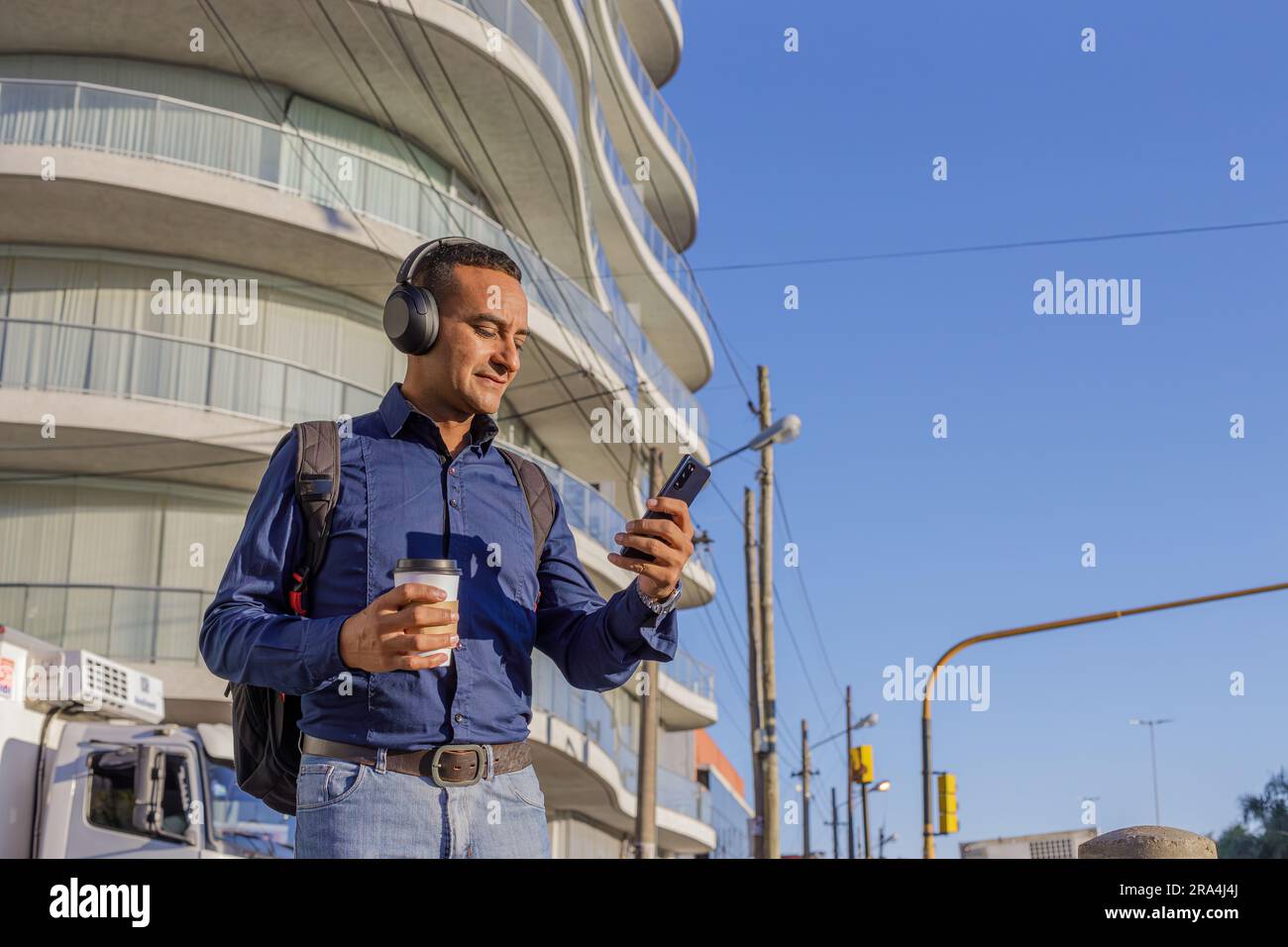 Young latin man with headphones looking at his mobile phone and with a paper cup with coffee in his hand in a city. Stock Photo