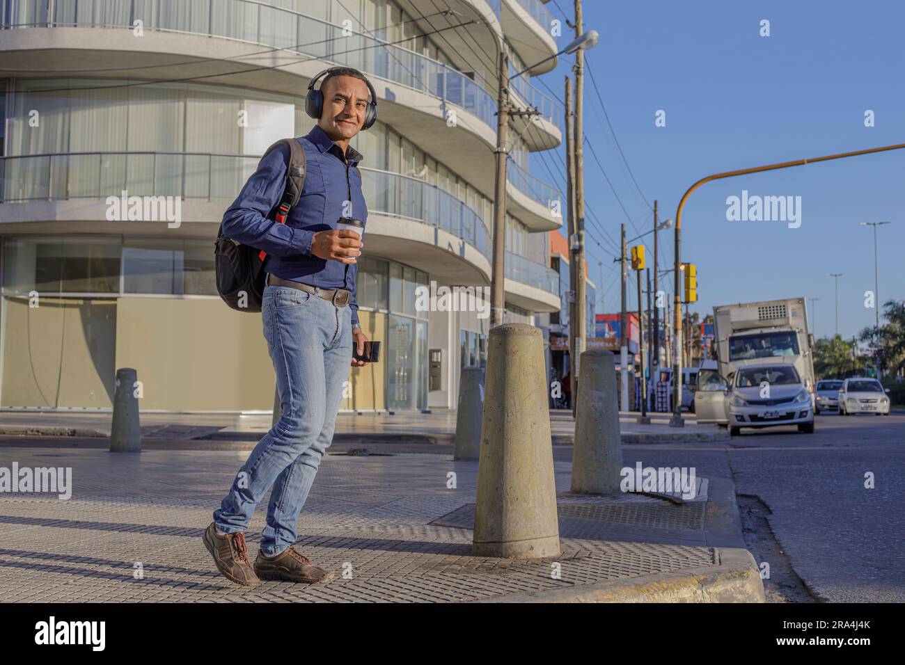 Young latin man with headphones and a paper cup with coffee in his hand walking through a city. Stock Photo