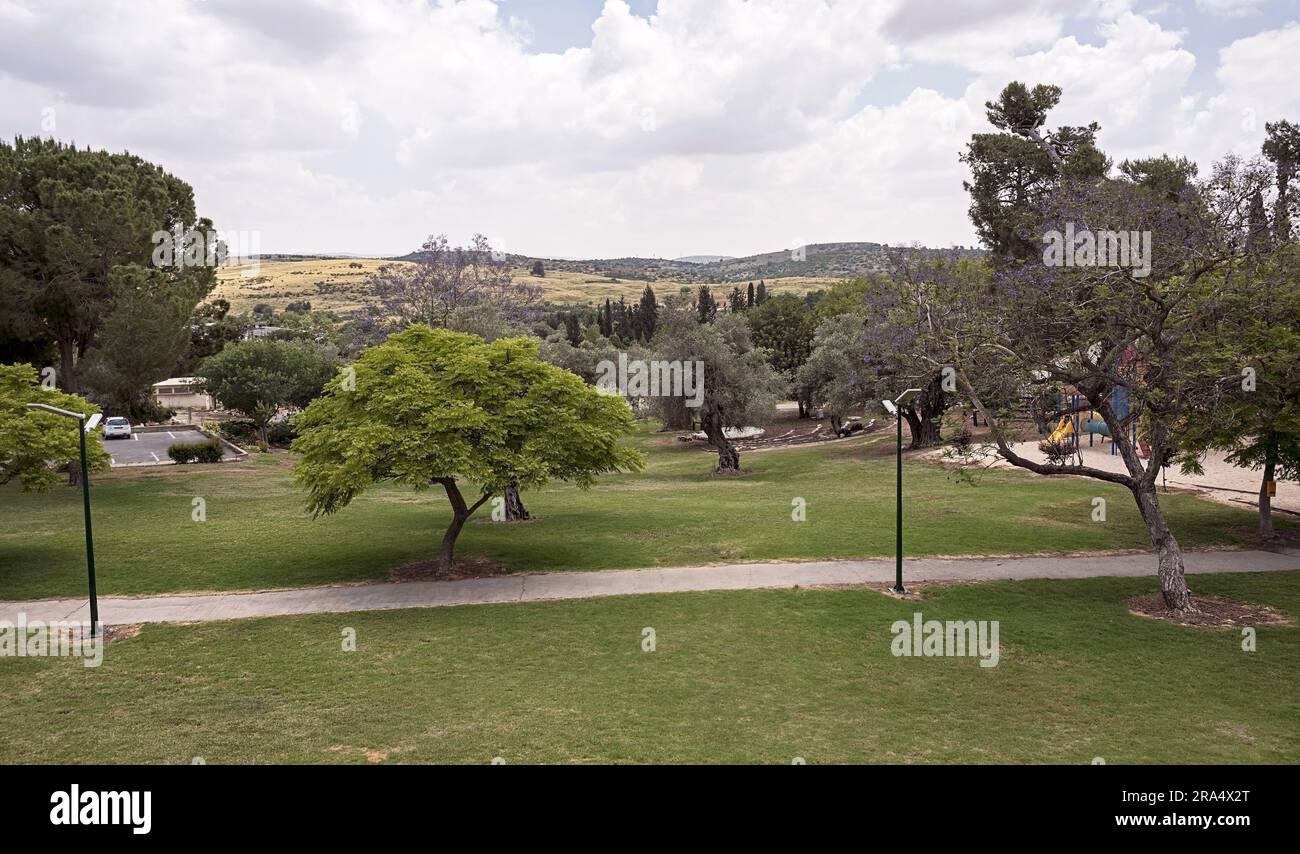 a wide walkway goes through an empty park with lawn and trees and a hillside a cloudy sky in the background Stock Photo