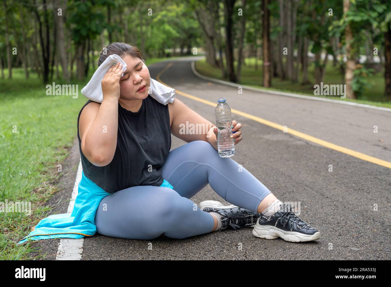 An overweight young woman wipping off her sweat from her forehead while holding water bottle and sitting down on the running track of a local park aft Stock Photo