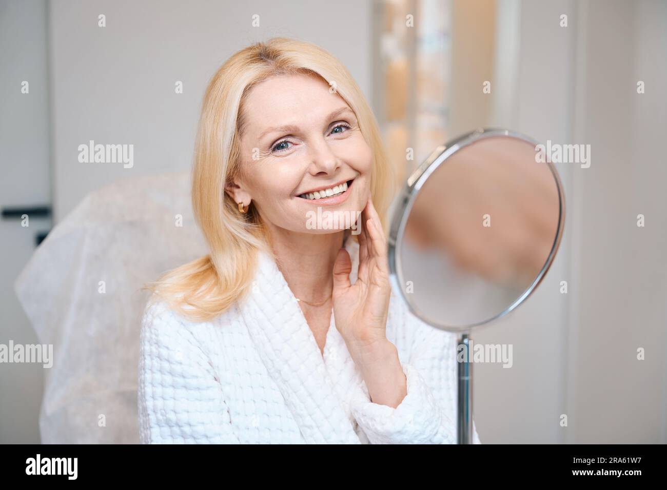 Smiling mature lady looking near mirror at cosmetologist office Stock Photo