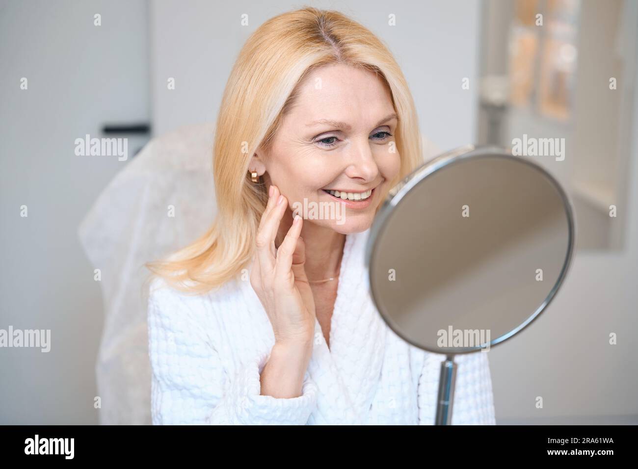Smiling mature lady looking in mirror at cosmetologist office Stock Photo