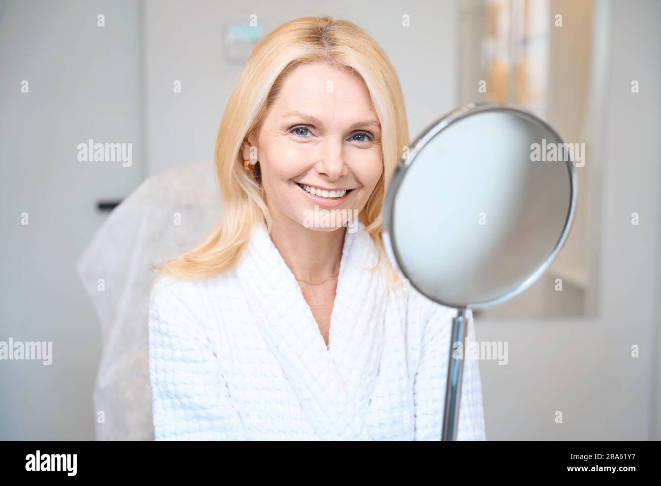 Smiling mature lady looking near mirror at cosmetologist office Stock Photo