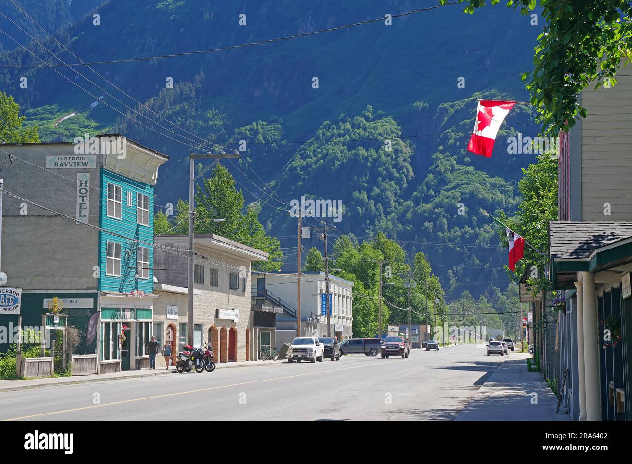Old wooden houses from the pioneer era in the main street of the village of Stewart, Canadian flag and green mountain landscapes, British Columbia Stock Photo