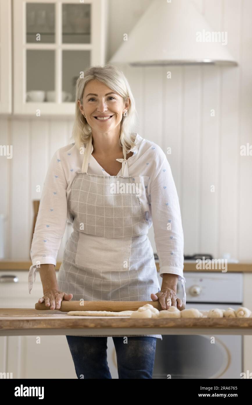 Happy pretty blonde mature baker woman in apron rolling dough Stock Photo