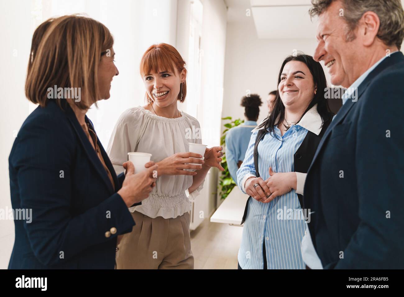 Group of colleagues in formal attire conversing and laughing near a window during an office break. Mix of senior and junior, set in a modern office. Stock Photo